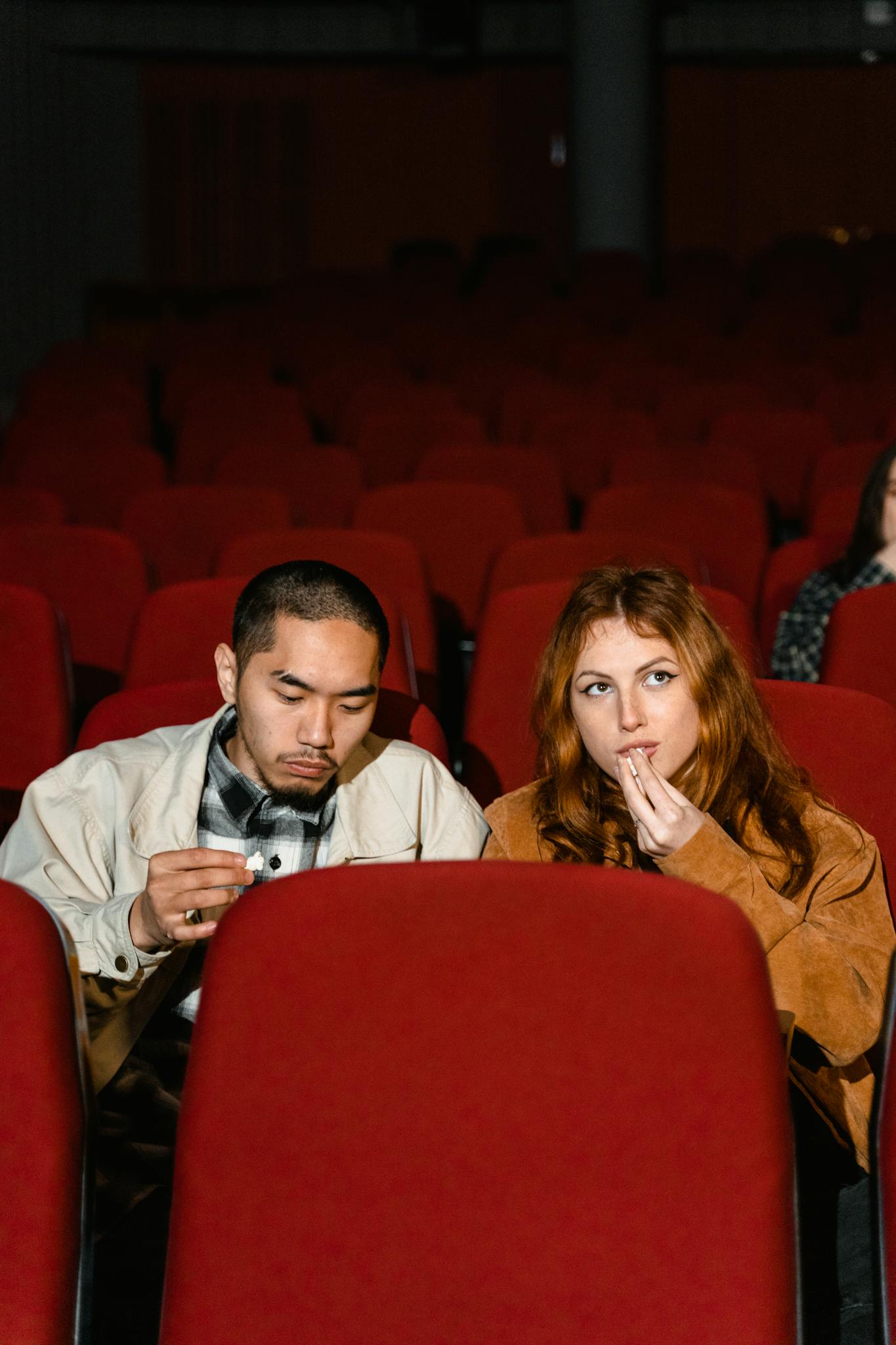 A couple seated in red cinema chairs enjoys a movie, sharing fun snacks.